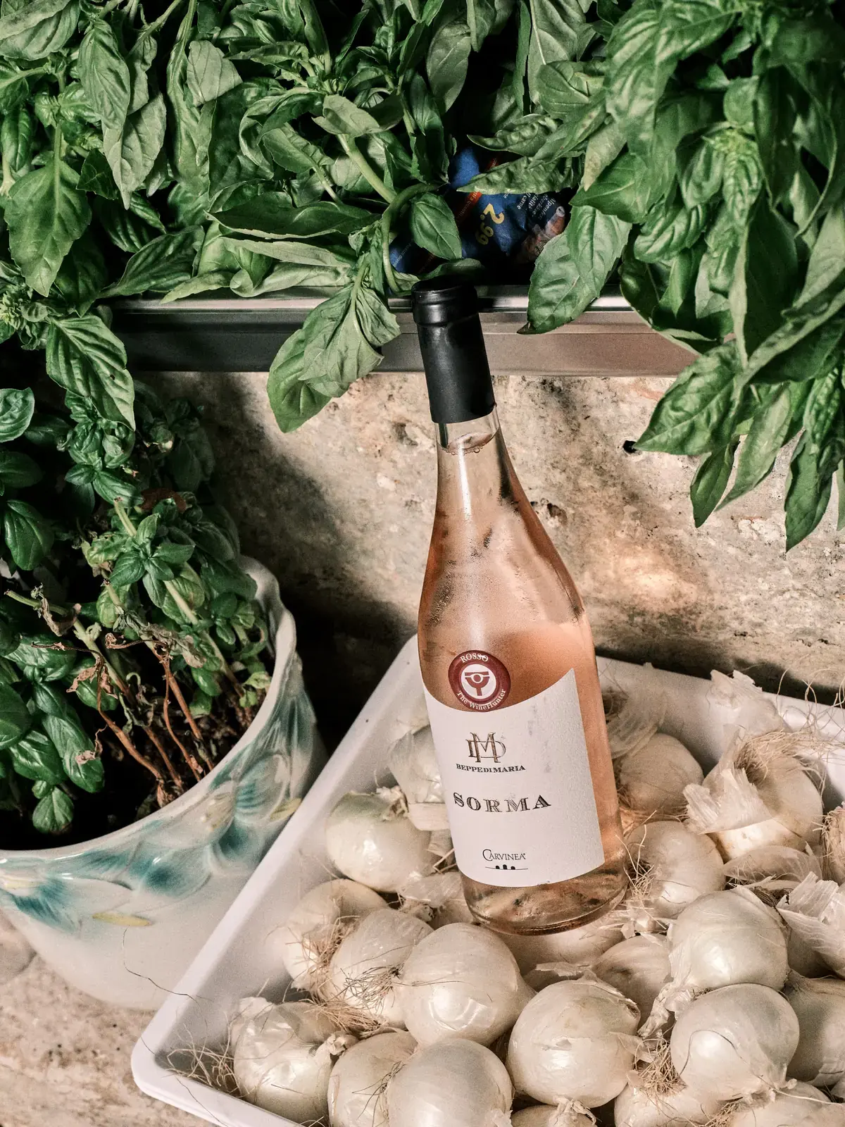 Una bottiglia di rosato pugliese appoggiata tra il basilico e le cipolle bianche in cucina