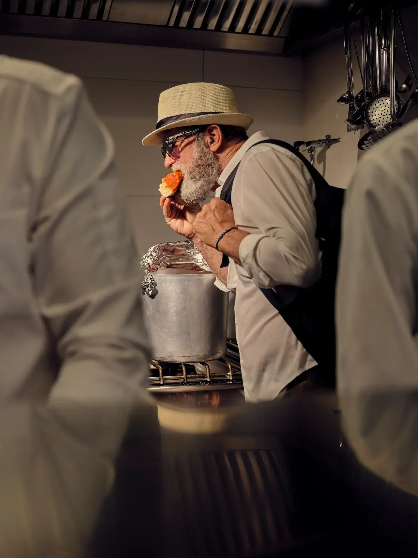 Nella cucina di NOS, visto tra le spalle di due cuochi: un uomo col cappello di paglia assaggia una bruschetta accanto alla pentola sul fuoco