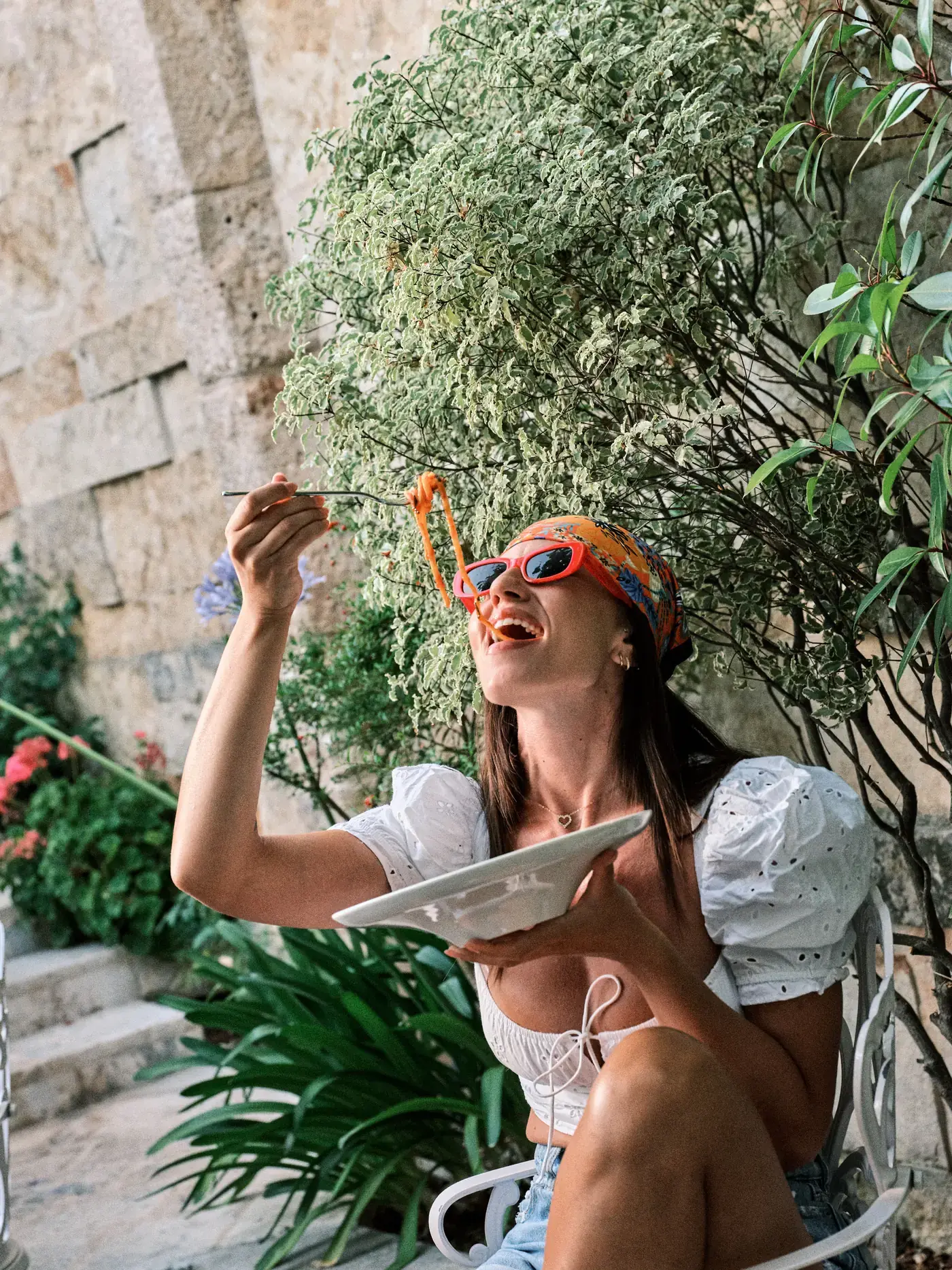 Una donna ride nel giardino di NOS ad Alberobello mentre solleva una forchettata di spaghetti al pomodoro, con la luce del giorno