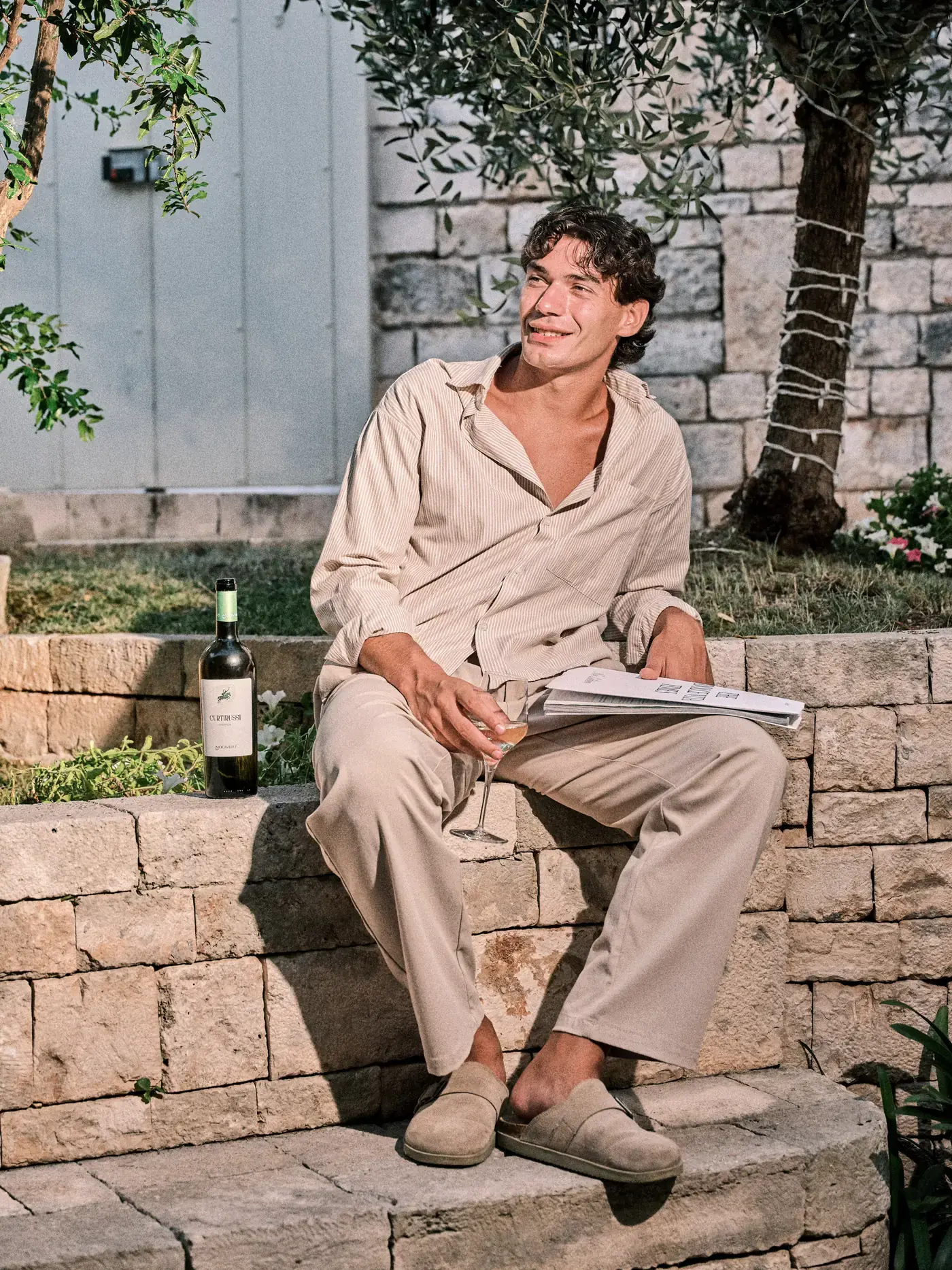 Un uomo seduto sul muretto di pietra sotto l'ulivo, nel giardino di NOS, con un calice di vino bianco e il giornale