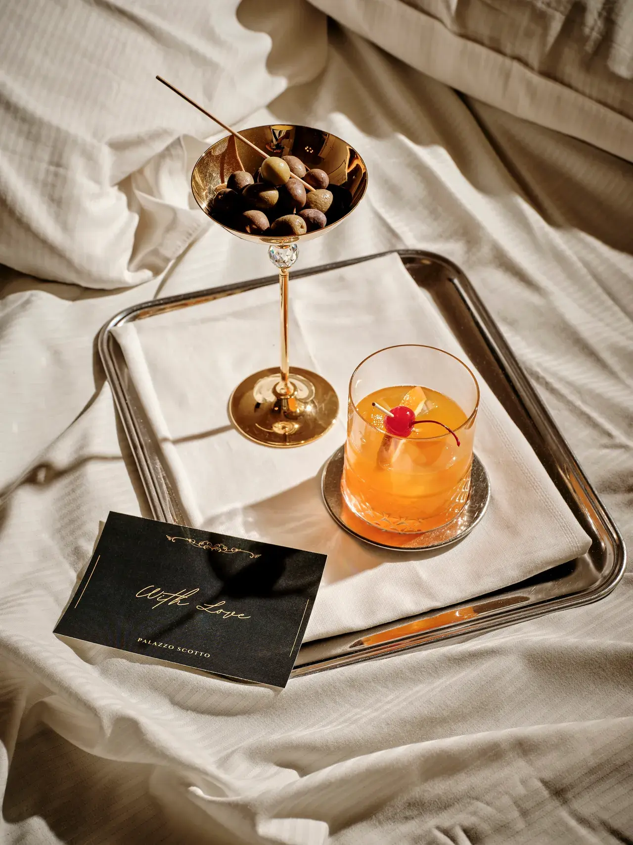 Cocktail arancio con coppa dorata su vassoio argento sul letto bianco, biglietto da visita Palazzo Scotto in scena