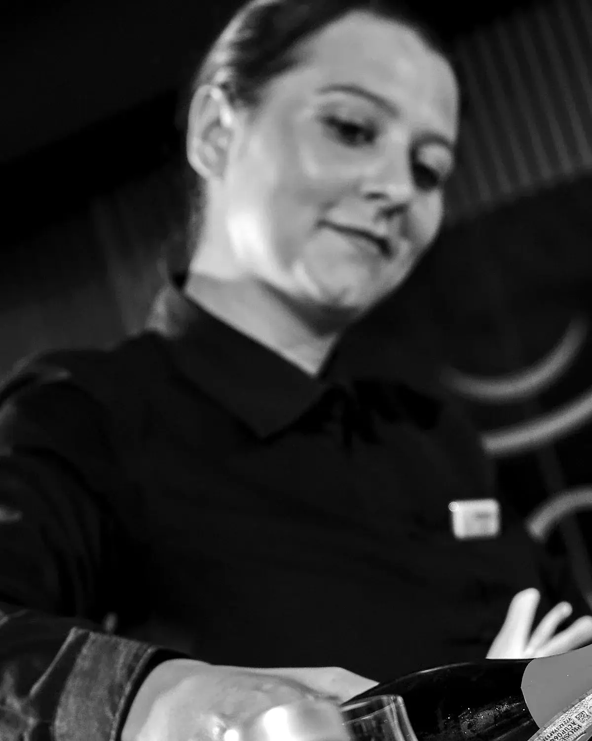 Federica, cameriera di sala, con i capelli raccolti e la divisa scura, versa del vino spumante nel calice di un ospite; intorno a lei alcuni bicchieri sul tavolo. Foto in bianco e nero.