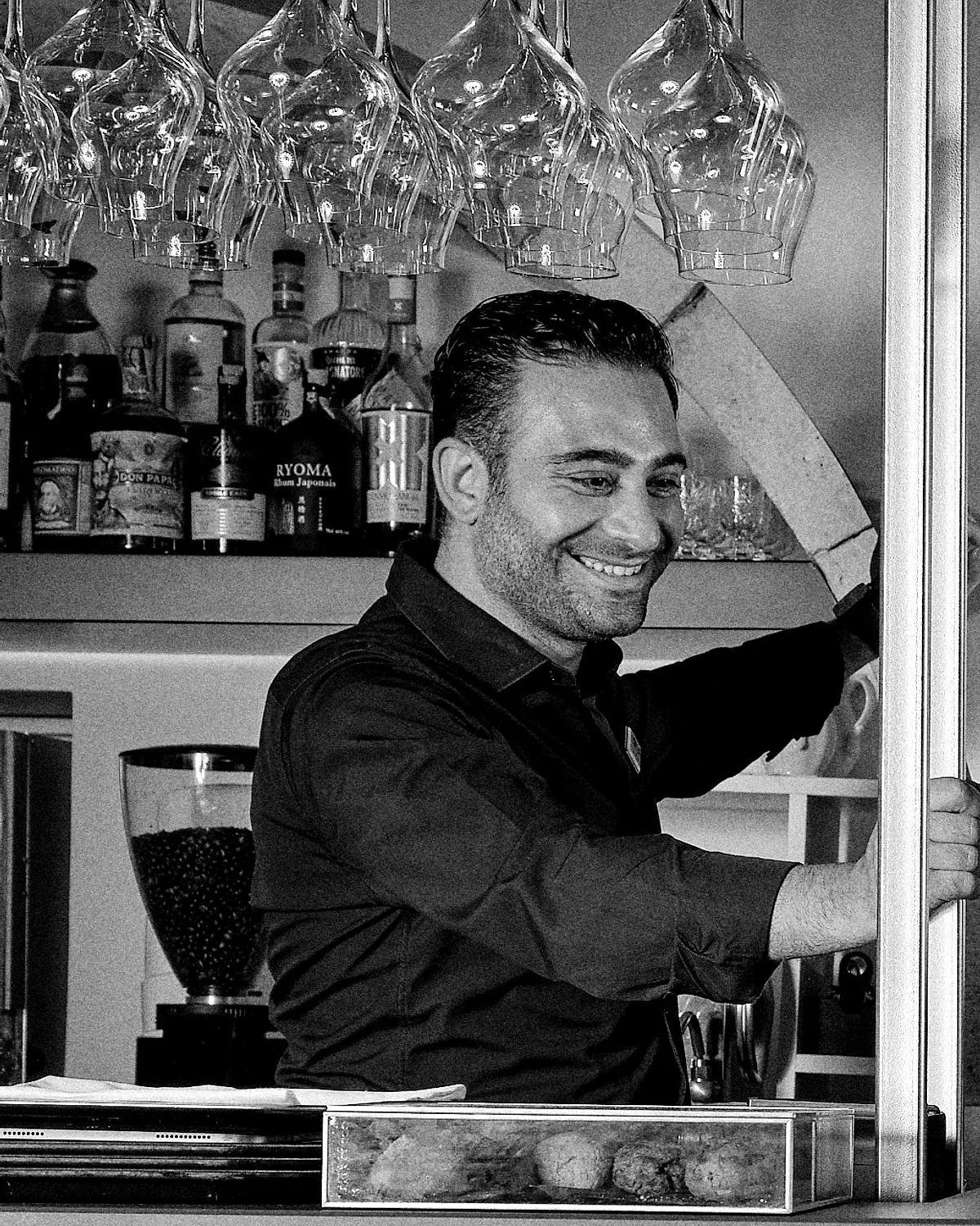Foto in bianco e nero. Un uomo dai capelli scuri, in camicia scura, sorride dietro al bancone mentre allunga la mano verso i bicchieri da vino appesi; alle spalle una mensola con bottiglie e, sul bancone, un vassoio di fritti.